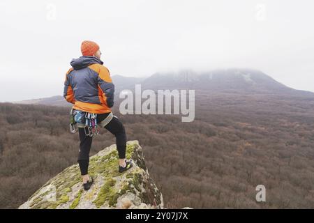 Hipster, un grimpeur dans une veste en duvet et une casquette tricotée est debout et médite en train de méditer en regardant le ponamramu automnal de la moun voisine Banque D'Images