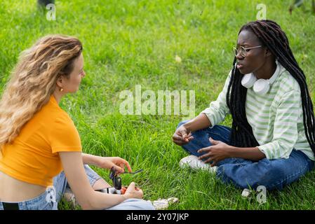 Deux jeunes femmes amies de différentes nationalités sont assises sur l'herbe dans le parc en été et parlent de sujets difficiles. Dialogue sur les problèmes sociaux. Le visage de la femme afro-américaine est tendu Banque D'Images