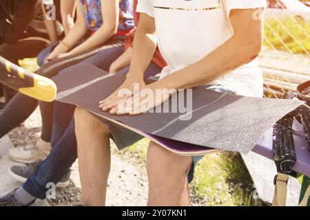 Un jeune garçon à genoux colle le griptape sur un skateboard en compagnie d'amis par temps ensoleillé. Préparation d'une planche à roulettes pour une compétition de skatepark Banque D'Images