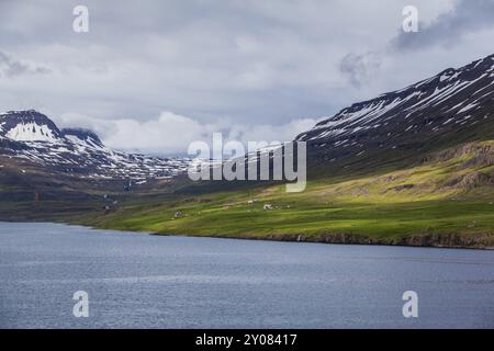 Quelques maisons dispersées se dressent sur une côte verdoyante en face des montagnes enneigées en Islande Banque D'Images