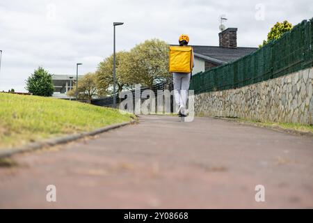 Fille de livraison de nourriture latine sur un scooter électrique, portant un sac à dos jaune et un casque, est vue de derrière alors qu'elle part dans une piste cyclable. L'image Banque D'Images