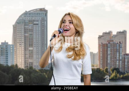 Portrait d'une jolie femme blonde émouvante chantant dans le micro. Bâtiments de zone résidentielle en arrière-plan. Banque D'Images