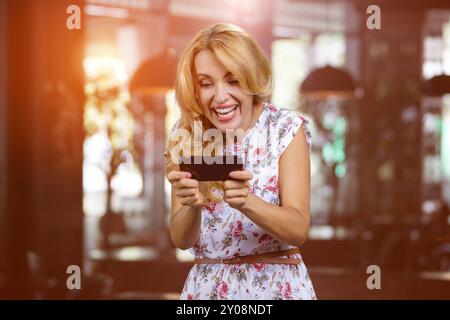 Heureuse femme mature blonde jouant au jeu vidéo en ligne sur son smartphone à l'intérieur. Intérieur de la maison en arrière-plan. Banque D'Images