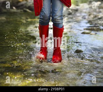 Enfant portant des bottes de pluie rouge sauter dans une rivière de montagne. Close up Banque D'Images