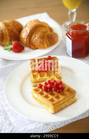 Gaufres aux fruits et de la confiture de groseille rouge sur une assiette blanche, croissants, jus d'orange sur le fond en bois Banque D'Images