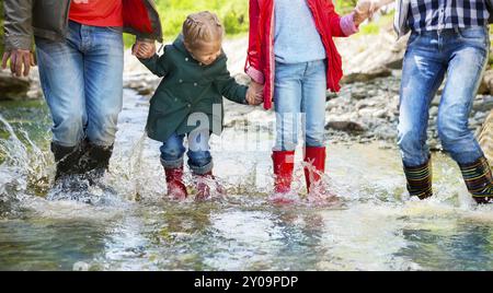 Famille heureuse avec deux enfants portant des bottes de pluie sauter dans une rivière de montagne Banque D'Images