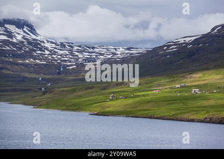 Quelques maisons dispersées se dressent sur une côte verdoyante en face des montagnes enneigées en Islande Banque D'Images