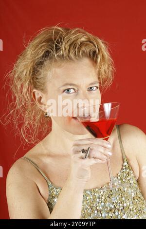 Portrait d'une belle femme blonde aux yeux bleus, portant un dessus de paillettes vertes, tenant une boisson rouge, sur un fond rouge. Flou de mise au point Banque D'Images