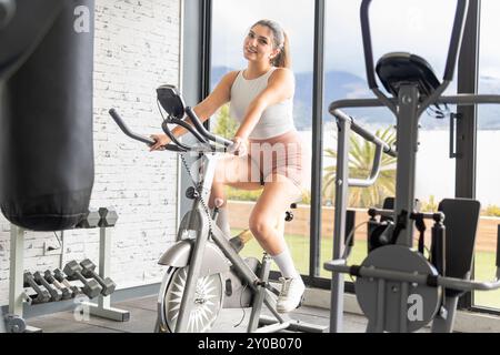 Jeune femme Latina regardant la caméra sur le vélo d'exercice dans la salle de gym au milieu d'autres machines d'exercice Banque D'Images