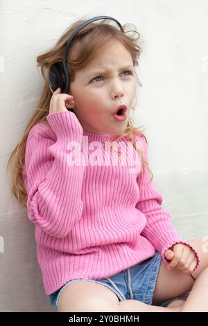 jolie petite fille d'âge préscolaire assise sur le sol appuyée contre le mur, écoutant de la musique dans des écouteurs et chantant Banque D'Images