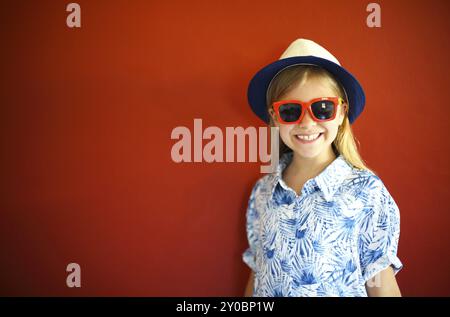 Très emothional enfant porter un chapeau et des lunettes de soleil sur fond rouge. Copie espace, lumière du jour Banque D'Images