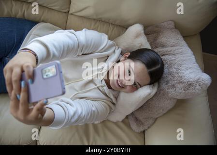 Vue aérienne d'une jeune femme caucasienne allongée sur un canapé, capturant un selfie. Moment de joie et d'expression de soi dans un environnement accueillant Banque D'Images