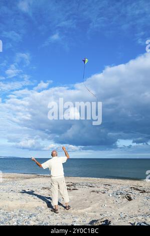 Un homme plus âgé et actif vole un cerf-volant sur une plage en face d'une mer calme et d'un paysage nuageux spectaculaire d'été. Un homme vole un cerf-volant sur une plage en face d'un calme s. Banque D'Images