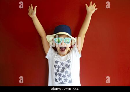 Très emothional enfant porter un chapeau et des lunettes de soleil sur fond rouge. Copie espace, lumière du jour Banque D'Images