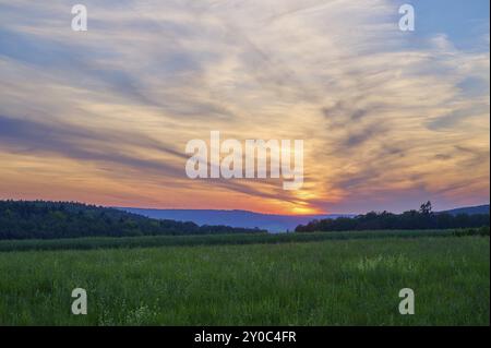 Champ vert avec des arbres en arrière-plan sous un coucher de soleil dramatiquement illuminé, le ciel est baigné de tons orange et bleu, Moenchberg, Mltenberg, S. Banque D'Images