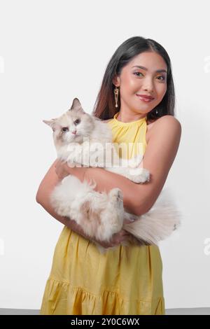 Souriant jeune femme asiatique en robe jaune tenant un chat ragdoll blanc sur fond blanc isolé. Concept de chaleur et d'affection. Banque D'Images