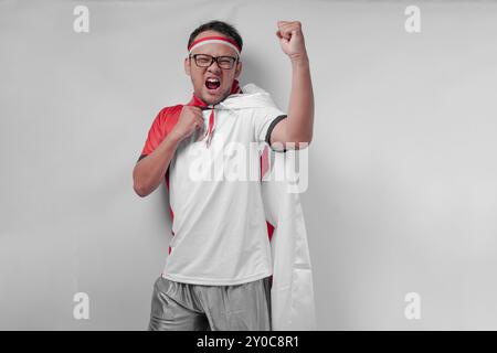 Jeune homme asiatique énergique portant un maillot avec drapeau drapé sur l'épaule levant la main dans les airs avec une expression heureuse. Jour de l'indépendance de l'Indonésie et Banque D'Images