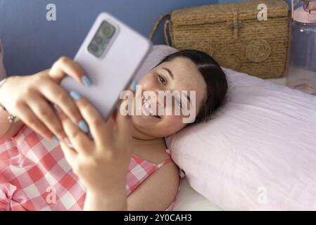 Belle et joyeuse jeune femme latino-américaine en pyjama, prenant un selfie avec son téléphone portable. Expression du bonheur et atmosphère chaleureuse, tarr Banque D'Images