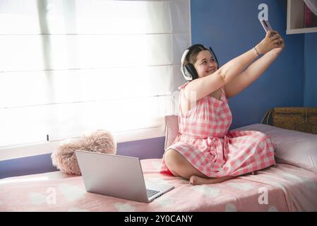 Une fille assise sur son lit, profitant d'un moment de loisir. Elle prend un selfie avec son téléphone, avec son ordinateur portable et un animal en peluche à proximité, tarragone Banque D'Images