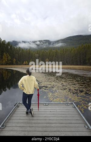 Une femme se tient debout sur un quai appuyé contre un parapluie rouge plié au lac Sinclair dans le nord de l'Idaho pendant l'automne Banque D'Images