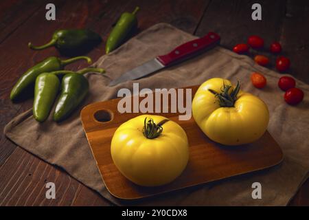 Deux tomates jaunes mûres sur une planche à découper, des poivrons verts épicés et quelques tomates cerises Banque D'Images