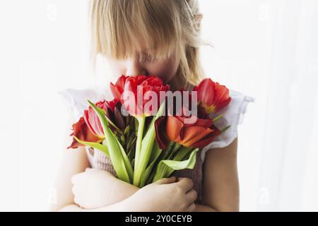 Petite fille souriante tenant et enniffant bouquet de fleurs de tulipe rouge. Concept de carte de voeux pour Pâques, Fête des mères, Journée internationale de la femme, S Banque D'Images