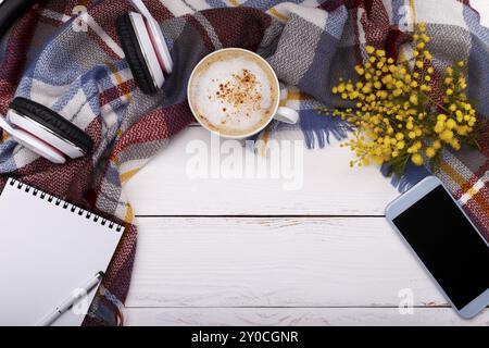 Cappucino de café chaud, couverture, fleurs de mimosa, bloc-notes, smartphone et écouteurs sur fond de bois ancien. Concept Relax saisonnier, pose à plat Banque D'Images