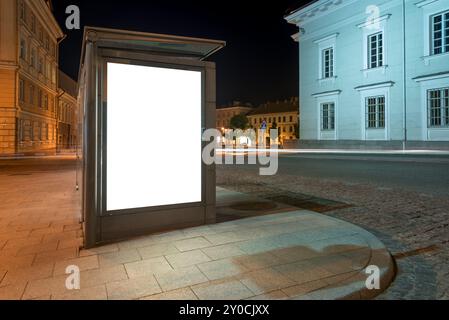 Maquette blanche vierge de panneau d'affichage vertical dans Un arrêt de bus la nuit. Une visionneuse publicitaire vide sur la place de la mairie Banque D'Images