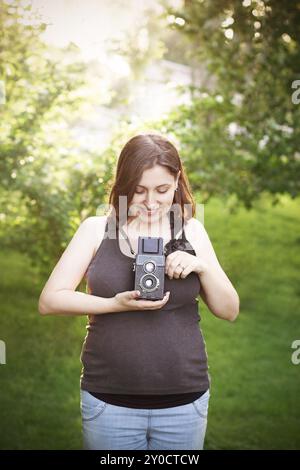 Charmante jeune femme enceinte dans une robe décontractée prenant des photos avec un appareil photo dans le parc vert en été ensoleillé Banque D'Images