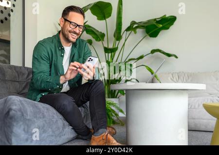 Pleine longueur de freelance utilisant des applications sur téléphone mobile tout en étant assis sur le canapé dans le bureau moderne Banque D'Images