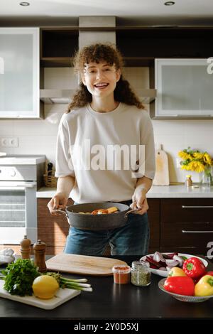 Femme joyeuse tenant une plaque de cuisson avec des légumes dans ses mains tout en se tenant dans la cuisine près de la table avec des produits biologiques frais Banque D'Images