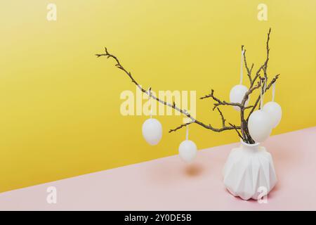 Joyeuses Pâques, concept du printemps. Branches d'arbre, brindilles de fleurs dans un vase blanc ou un pot avec des œufs suspendus. Fond de table jaune et rose avec Banque D'Images