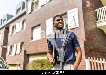 Fier jeune homme d'affaires africain avec des vêtements décontractés portant un ordinateur portable regardant loin debout dans la rue dans un quartier résidentiel Banque D'Images