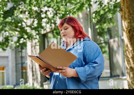 Une femme de grande taille avec les cheveux rouge vif lit un dossier à l'extérieur. Banque D'Images