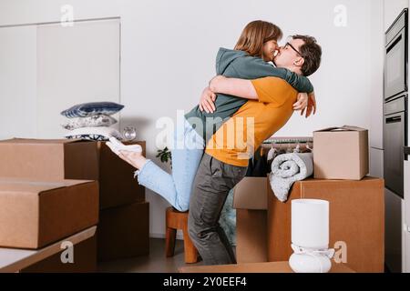 Le couple s'embrasse et se soulève dans une nouvelle maison, entouré de boîtes mobiles, célébrant avec joie et rire Banque D'Images