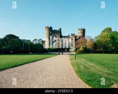 Deux personnes marchent le long d'un chemin de gravier menant à un château impressionnant, encadré par des arbres vibrants et une verdure lumineuse par une journée claire et ensoleillée. Banque D'Images