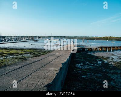 Le front de mer offre une large vue sur les bateaux amarrés à marée basse, avec la surface calme reflétant le ciel bleu et les collines lointaines sous la lumière du soleil Banque D'Images