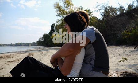 Couple romantique embrasse pendant assis sur le rondin de bois sur la plage. Paire heureuse admirant une mer et un paysage. Amoureux passant du temps ensemble à la station Banque D'Images