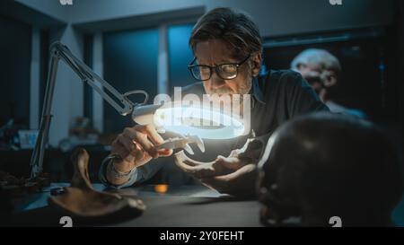 Archéologue mature prend des mesures de l'os fossile à l'aide d'un outil de mesure professionnel, étudie les restes de l'homme préhistorique sous la lampe grossissante. Le scientifique travaille dans un laboratoire archéologique la nuit. Banque D'Images