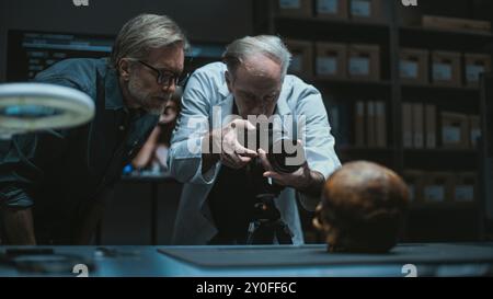 Deux archéologues parlent et prennent des photos de restes fossiles du crâne humain préhistorique en utilisant un appareil photo professionnel sur trépied. Les scientifiques travaillent dans un laboratoire archéologique moderne. Concept d'archéologie. Banque D'Images