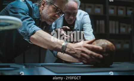 Deux archéologues parlent et prennent des photos de restes fossiles du crâne humain préhistorique en utilisant un appareil photo professionnel sur trépied. Les scientifiques travaillent dans un laboratoire archéologique moderne. Concept d'archéologie. Banque D'Images