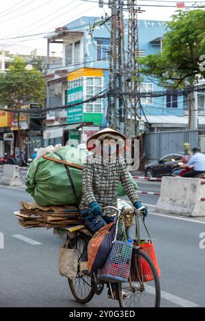 Nha Trang, Vietnam - 22 avril 2024 : une femme âgée vietnamienne en vêtements fermés traditionnels et chapeau porte des ordures sur un vieux vélo le long de la ville Banque D'Images