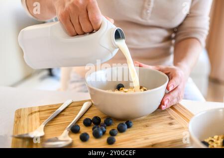 Une fille met du lait aux flocons d'avoine avec des myrtilles dans un bol pour un délicieux petit déjeuner de céréales dans la cuisine. Femme préparant du granola avec des baies et du latte pour un repas diététique du matin Banque D'Images