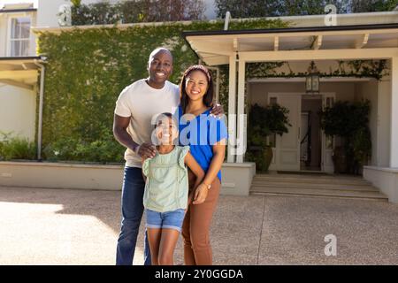 Famille souriante de trois avec fille debout ensemble à l'extérieur de leur maison, profitant du temps Banque D'Images