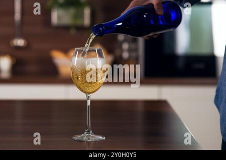 Homme méconnaissable versé dans un verre de vin blanc dans des bulles avec une éclaboussure sur la table dans la cuisine, gros plan Banque D'Images