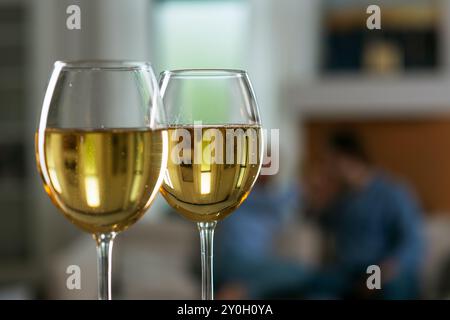 Vin blanc dans des verres avec une photo floue d'un couple aimant assis sur un canapé dans le salon en arrière-plan Banque D'Images