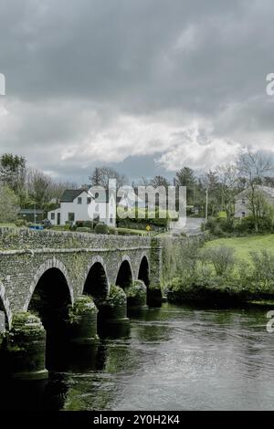 Un pont de pierre aux multiples arches traverse une rivière, avec une route menant à un petit village de l'autre côté. Le ciel est couvert. Pont sur le Ri Banque D'Images