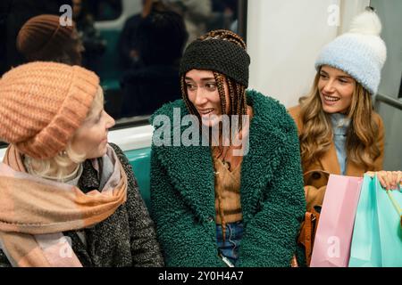 Groupe de jeunes amies diverses parlant et riant dans les transports publics. Porter des tenues hivernales tendance et transporter des sacs à provisions, mettre en valeur fr Banque D'Images