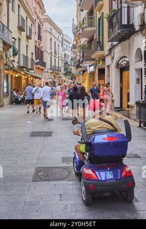 Sitges, Barcelone-01 septembre 2024 : individuel sur un fauteuil roulant électrique visitant une rue animée avec des magasins et des piétons à Sitges, Barcelone. Le i Banque D'Images