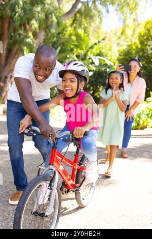 Enseigner à la fille à faire du vélo, père guidant tandis que mère et sœur regardent Banque D'Images